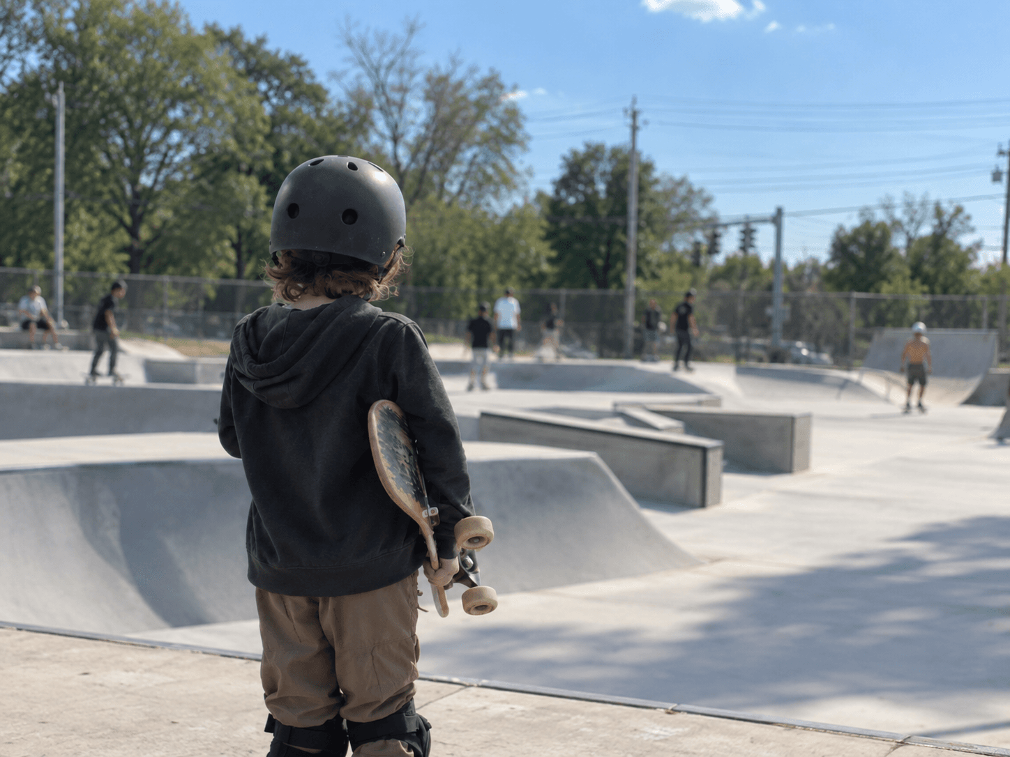 Young kid holding a skateboard and looking out at a busy skatepark
